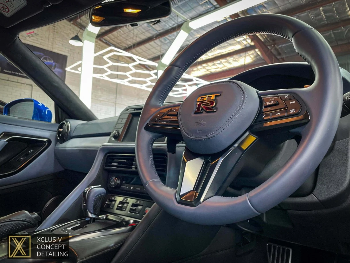 Close-up of a Nissan GT-R steering wheel and dashboard inside a modern garage, with ceiling lights reflected on the car’s interior surfaces. A logo for XCLUSIV Concept Detailing is visible in the lower left corner.