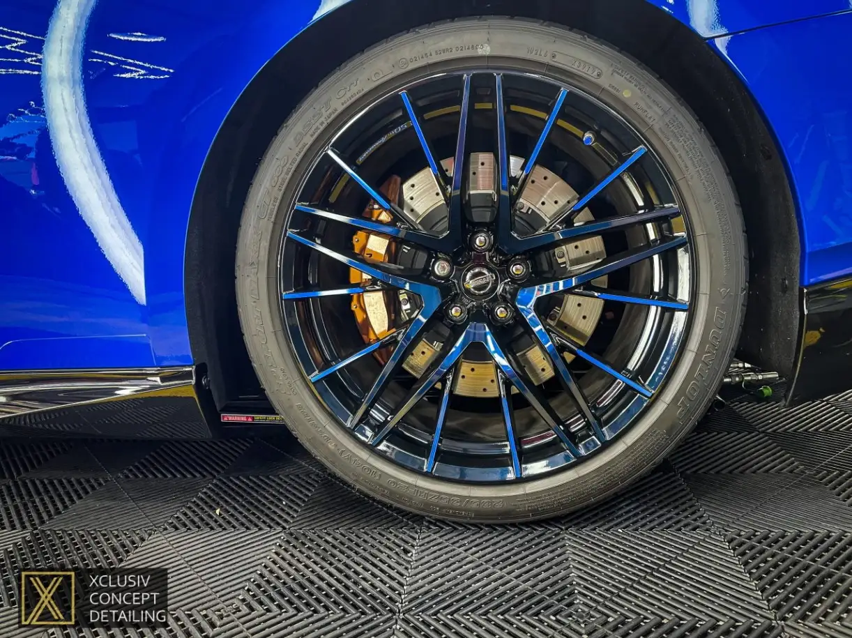 Close-up of a black car wheel with blue accents, showing detailed brake discs and calipers. The Nissan GTR has a blue body is partly visible, and the floor has a textured black pattern with a logo of Xclusiv in the corner.