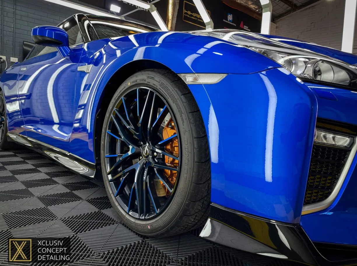 Close-up view of a shiny blue GTR Nissan front wheel and fender, showcasing black alloy wheels, orange brake calipers, and polished bodywork, parked indoors on a checkered floor.