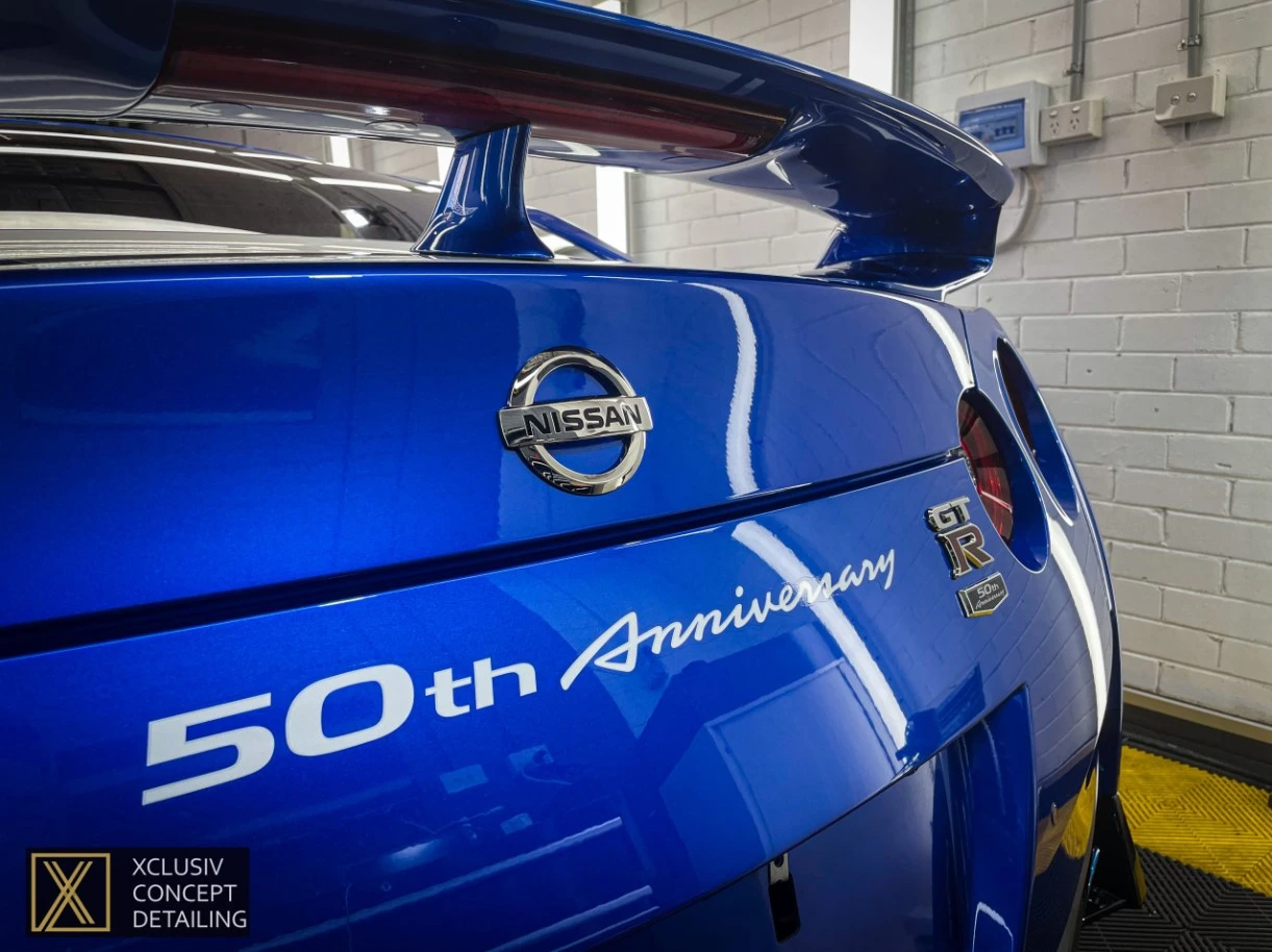 Close-up of the rear of a blue Nissan GT-R 50th Anniversary edition car, showcasing its badge, special edition emblem, and rear spoiler inside a garage with white brick walls.