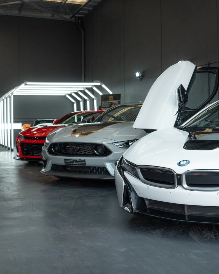 A row of three sports cars, white BMW with a lifted scissor door, grey Ford Mustang, and red Chevrolet Camaro, parked inside a our new modern garage with a lit geometric light structure in the background.