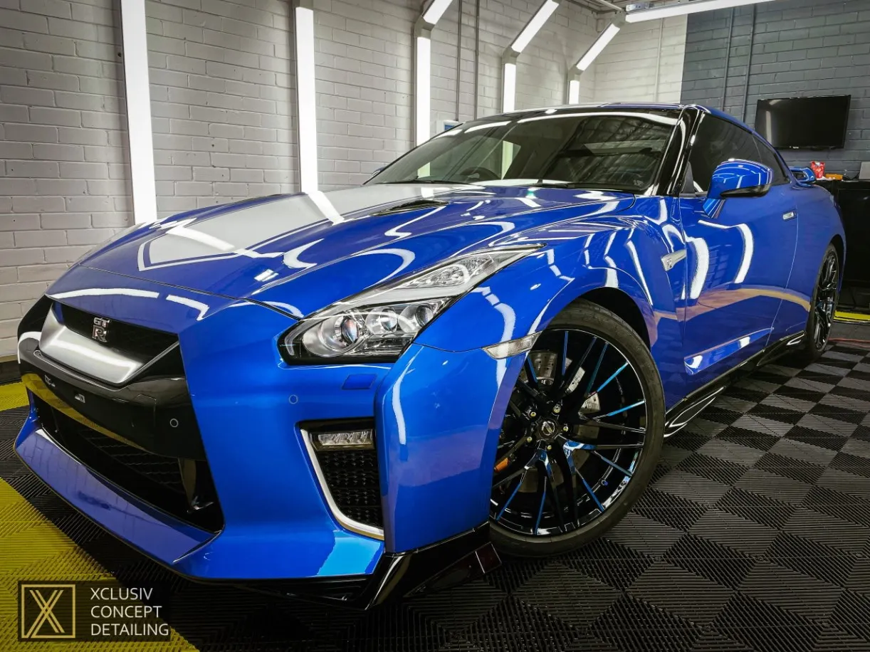 A shiny blue Nissan GTR sports car with black and chrome accents is parked in a brightly lit garage with white walls and checkered flooring. The car is highly polished, reflecting the lights above.