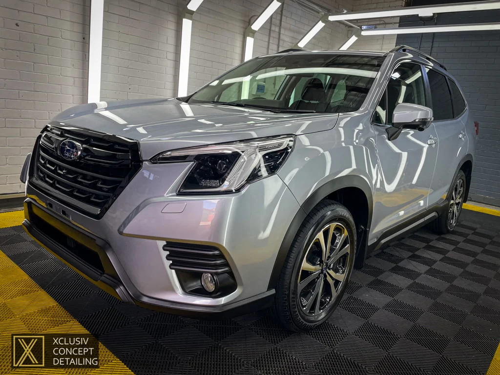 A silver Subaru Forester SUV parked indoors on a black and yellow checkered floor, with bright overhead lights reflecting on its polished surface. A detailing studios logo is visible in the bottom left corner.