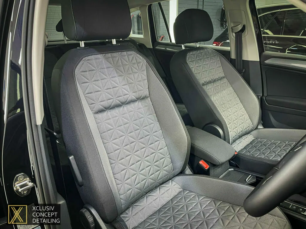 The image shows the clean, detailed interior of VW Tiguan featuring two front seats with grey, diamond-patterned upholstery. The car door is open, and a logo for Xclusiv Concept Detailing is visible in the bottom left corner.